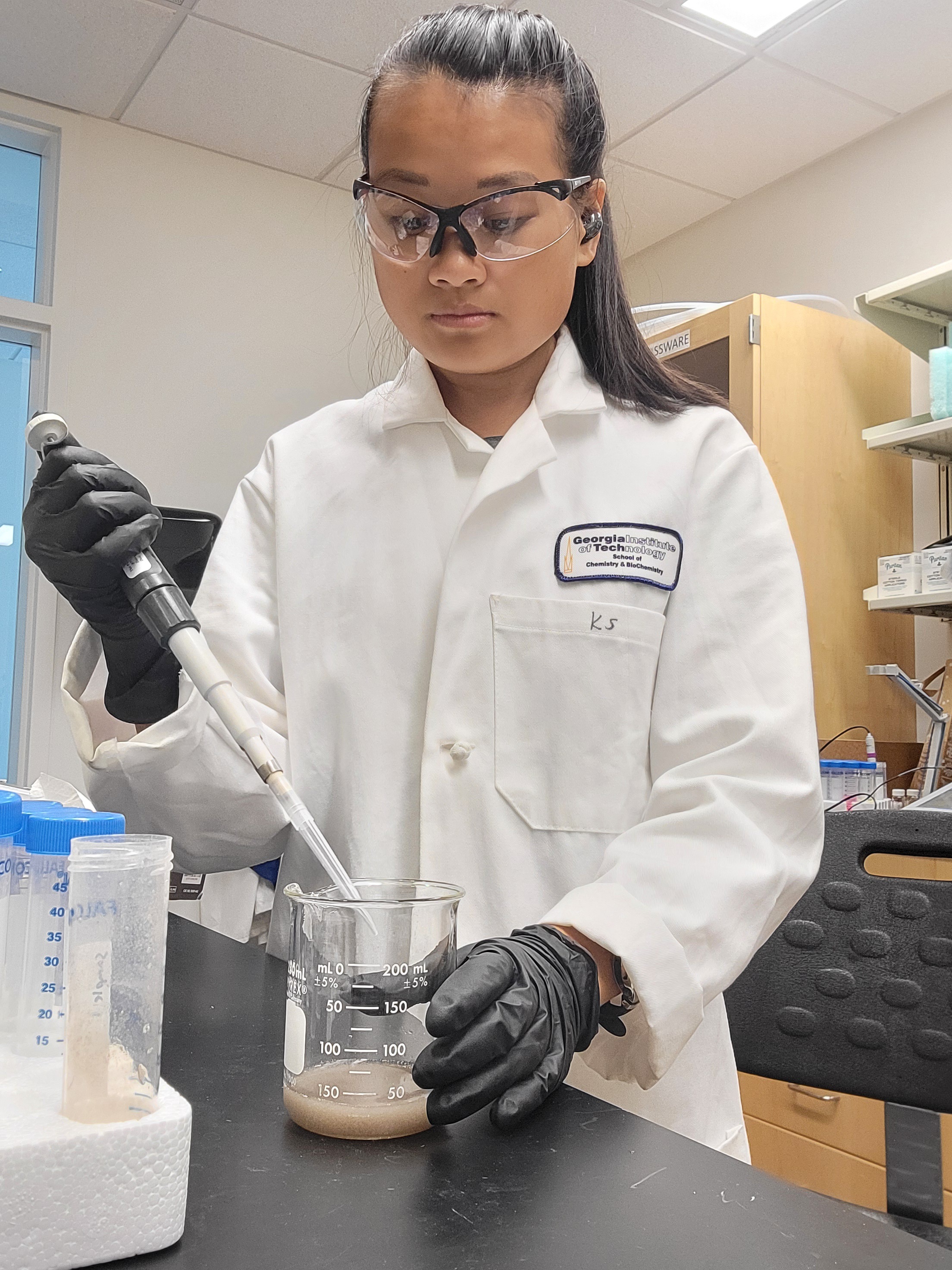Katie, wearing a lab coat and protective glasses, uses a pipette with a liquid in a beaker.