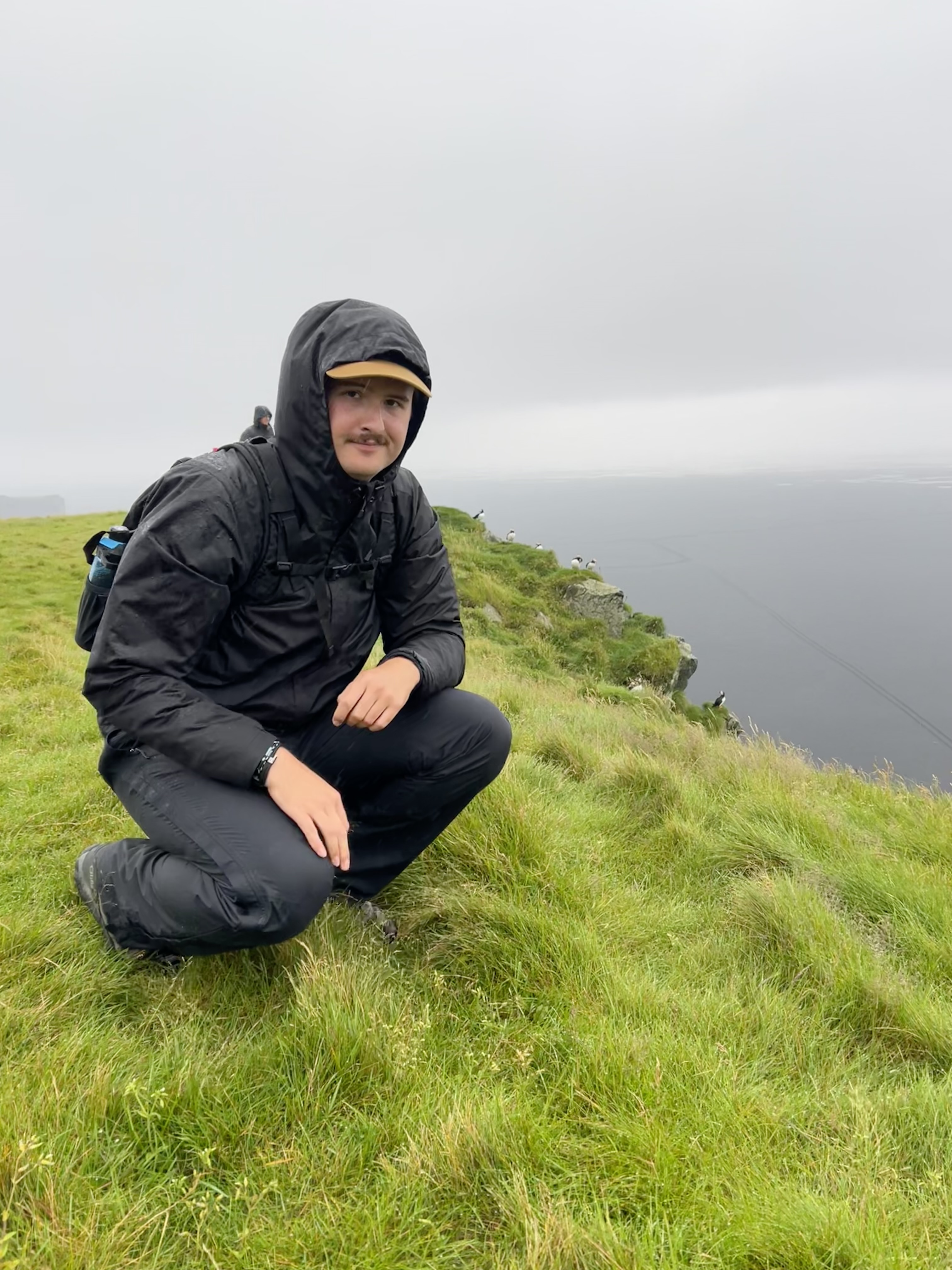 Eli poses in a crouch, wearing a hooded rain jacket, on a grassy cliff with a gray sea behind him.