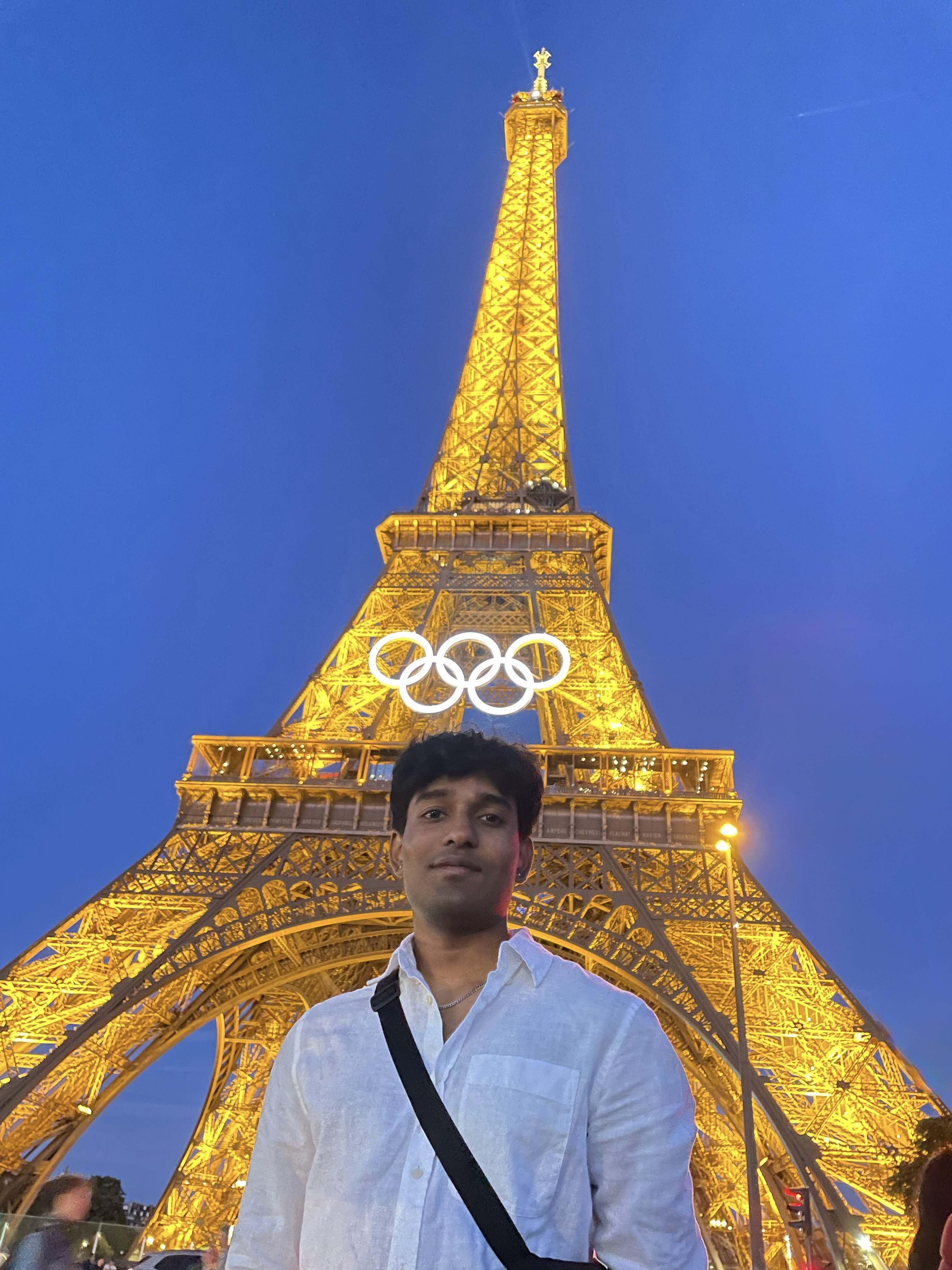 Pranay smiles at the camera with the Eiffel Tower displaying the Olympic Rings illuminated behind him.
